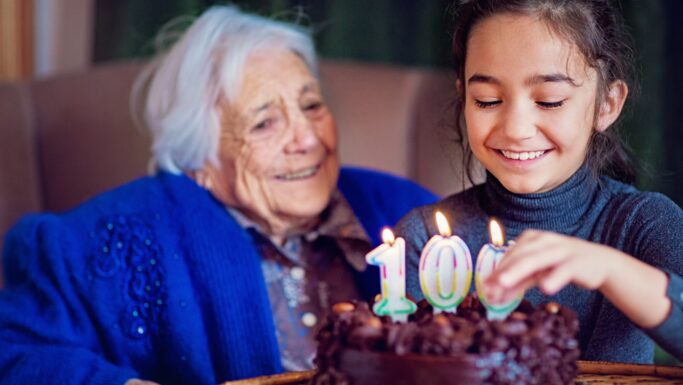 En ung pige tænder lys på en chokoladekage med tallet 100, mens en ældre kvinde i en blå sweater smiler og ser på, mens de fejrer hendes 100-års fødselsdag og lange levetid sammen.