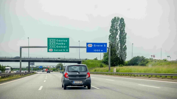 En sort bil kører på en motorvej i Danmark mod vejskilte til Odense, Røby, Gedser, Solrød N og Greve S. Andre biler og en viadukt er synlige forude, mens bilisterne bliver mindet om at holde til højre. Himlen er overskyet.