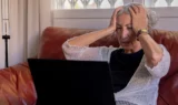 En ældre kvinde med gråt hår sidder i en rød sofa, ser stresset ud og holder hovedet i hænderne, mens hun stirrer på en bærbar skærm - muligvis bekymret for digital svindel. Lyset fra et vindue gør baggrunden lysere.
