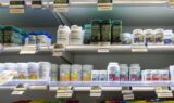 Flasker med vitaminer og kosttilskud står pænt arrangeret på hylderne i en butik, hver med elektroniske prisskilte under. Etiketterne har farverige designs og tekst, der viser forskellige mærker.