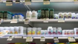 Flasker med vitaminer og kosttilskud står pænt arrangeret på hylderne i en butik, hver med elektroniske prisskilte under. Etiketterne har farverige designs og tekst, der viser forskellige mærker.