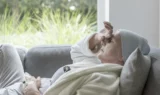 En ældre person med tørklæde og morgenkåbe ligger på en sofa og ser træt ud med en hånd på panden og et synligt IV-kateter - hvilket indfanger den stille kamp bag mange kræftsager - ved siden af et lyst vindue med grønne planter udenfor.