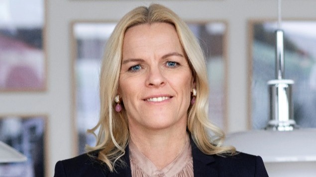 Ældreminister Mette Kierkgaard, med langt blondt hår, iført mørk blazer og lys bluse, smiler til kameraet. Den let slørede baggrund viser indrammede billeder og hvide hængelamper.