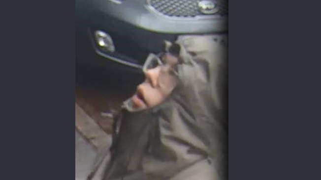 En person i grå hættejakke og briller, muligvis efterlyser mand i en svindel-sag, kigger opad med let åben mund og står i nærheden af en mørk bil på et vådt fortov.