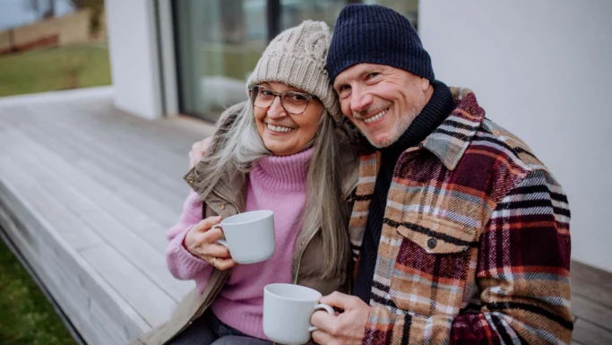 Et ældre par i varmt tøj og strikhuer sidder på en veranda, smiler og holder hvide krus og overvejer at samle pensionsordninger.