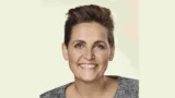 En person med kort, mørkt hår og lys hud smiler til kameraet. Vedkommende er iført en grå, struktureret top og små ørestikker, og baggrunden er ensfarvet og lys og minder om et foto fra valgkamp med Pia Olsen Dyhr.