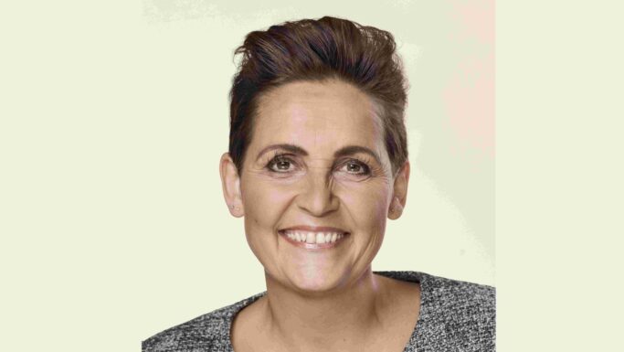 En person med kort, mørkt hår og lys hud smiler til kameraet. Vedkommende er iført en grå, struktureret top og små ørestikker, og baggrunden er ensfarvet og lys og minder om et foto fra valgkamp med Pia Olsen Dyhr.