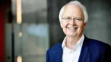 En ældre mand med hvidt hår og briller smiler varmt iført blå habitjakke og hvid skjorte. Baggrunden er blødt sløret med neutrale farver, der antyder den indbydende tilstedeværelse af en professor med viden om kostvaner.