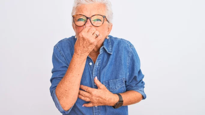 En ældre kvinde med briller og denimskjorte holder sig for næsen med den ene hånd og for maven med den anden og laver et sjovt ansigt, som om hun reagerer på mad med ildelugtende prutter.