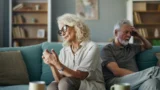 En ældre kvinde og en mand sidder i en sofa og ser oprørte og fjerne ud, mens de vender sig væk fra hinanden - en scene, der afspejler grå skilsmisser. Kvinden virker eftertænksom og trist, manden holder sig for panden. Det svagt oplyste rum føles både hyggeligt og anspændt.