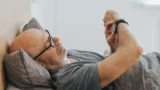 En ældre mand med briller ligger i sengen og hviler hovedet på en grå pude, mens han tjekker sit søvntracker-smartwatch. Han ser afslappet ud og er iført en grå t-shirt.