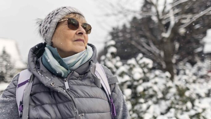 En ældre kvinde med solbriller, en grå strikhue og en puffy jakke står udenfor i et snedækket landskab og kigger op. Sneen falder, mens hun øver sig i at orientere sig i sneen - at navigere i vinterverdenen, som mange svagsynede gør.