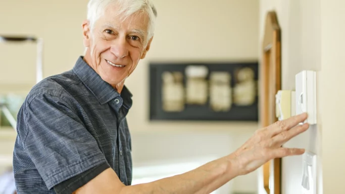 Ældre mand med gråt hår og kortærmet skjorte smiler, mens han justerer en termostat, glad for sin lavere elregningen i et godt oplyst hjem.