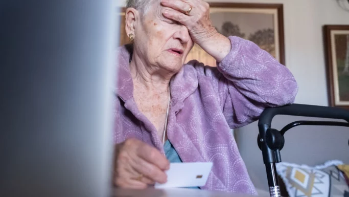 En ældre person i en lilla kappe sidder ved et bord, holder et kort og hviler hovedet i hånden og ser bekymret ud - muligvis bekymret for svindel eller falske sms'er. Et gangstativ og husholdningsartikler er synlige i baggrunden.