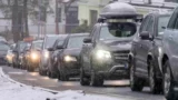 En række biler kører langsomt i snevejr under skolernes vinterferie med forlygterne tændt. Sneen dækker jorden og nogle biltage, hvilket skaber vinterlige og glatte vejforhold.
