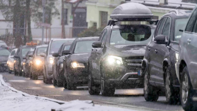 En række biler kører langsomt i snevejr under skolernes vinterferie med forlygterne tændt. Sneen dækker jorden og nogle biltage, hvilket skaber vinterlige og glatte vejforhold.