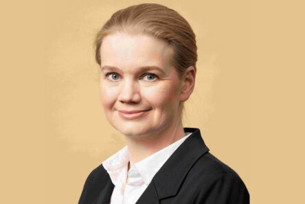 En kvinde med lys hud og tilbagestrøget blondt hår, iført en hvid skjorte med krave og en sort blazer, smiler let foran en almindelig beige baggrund og udstråler selvtillid og løftet om tryg alderdom.
