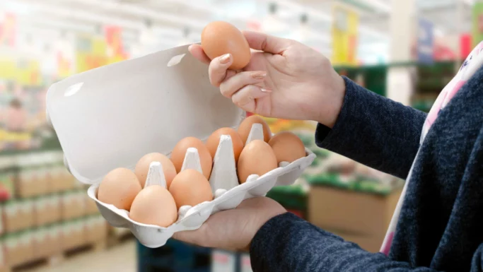 En person holder en karton med brunt æg fra Lidl i den ene hånd og samler et enkelt æg op med den anden inde i en købmandsbutik. Hylder og farverige skilte er synlige i den slørede baggrund.