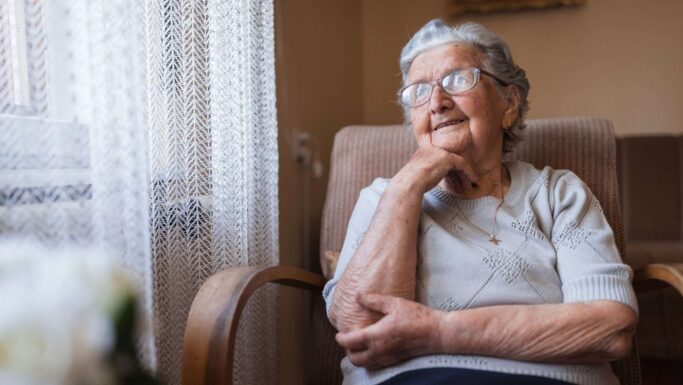 En ældre kvinde med briller sidder i en lænestol ved et vindue og smiler eftertænksomt. Hun hviler hagen på sin hånd og kigger ud, måske reflekterer hun over valgkampen, og hvordan ældre er blandt vælgernes emner. Blødt naturligt lys fylder rummet.