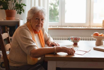 En ældre kvinde med gråt hår og orange tørklæde sidder ved et træbord i sin hyggelige, solbeskinnede plejebolig ved siden af en tekop, en skål med frugt og bøger og kigger eftertænksomt på kameraet.