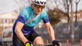 En ældre mand iført hjelm, solbriller og blå cykeltrøje cykler udendørs på en overskyet dag. Træer og bygninger er synlige i den slørede baggrund, mens han styrker sine muskler med hver kort træning.