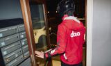 En person i rød dao-jakke og sort hjelm åbner en dør for at gå ud om natten. Langs væggen til venstre står postkasser, som minder om sikkerhed og advarer mod falske dao-beskeder.