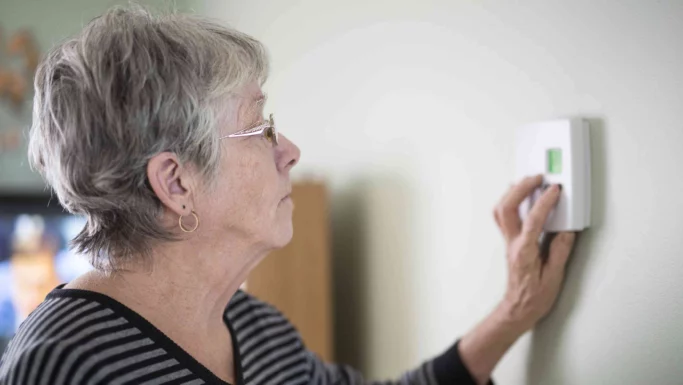 En ældre kvinde med kort gråt hår og briller, iført en sort og hvid stribet skjorte, justerer en digital termostat på væggen, mens hun overvejer, hvilken elaftale der er bedst, inspireret af sin seneste elaftale-test.