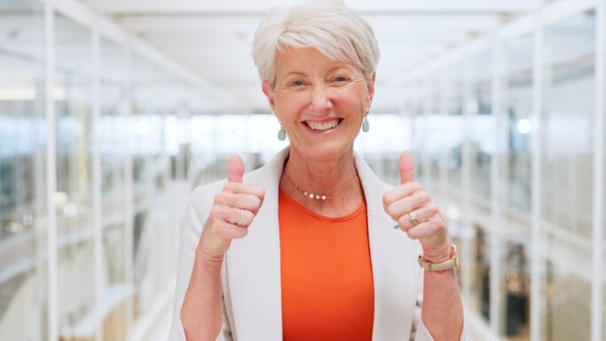 Smilende ældre kvinde med kort hvidt hår, iført orange top og hvid blazer, giver to tommelfingre op og fejrer et ekstra år på jobbet i lyse, moderne omgivelser med glasvægge.