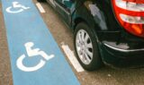 En sort bil er parkeret ved siden af en blå handicapparkeringsplads, som er markeret med et hvidt kørestolssymbol på fortovet.