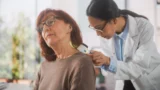 En kvindelig læge undersøger en ældre kvindes hals med et dermatoskop i en lys lægeklinik og tjekker for tegn eller symptomer, mens patienten kigger opad, og lægen fokuserer nøje på huden.
