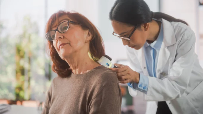 En kvindelig læge undersøger en ældre kvindes hals med et dermatoskop i en lys lægeklinik og tjekker for tegn eller symptomer, mens patienten kigger opad, og lægen fokuserer nøje på huden.