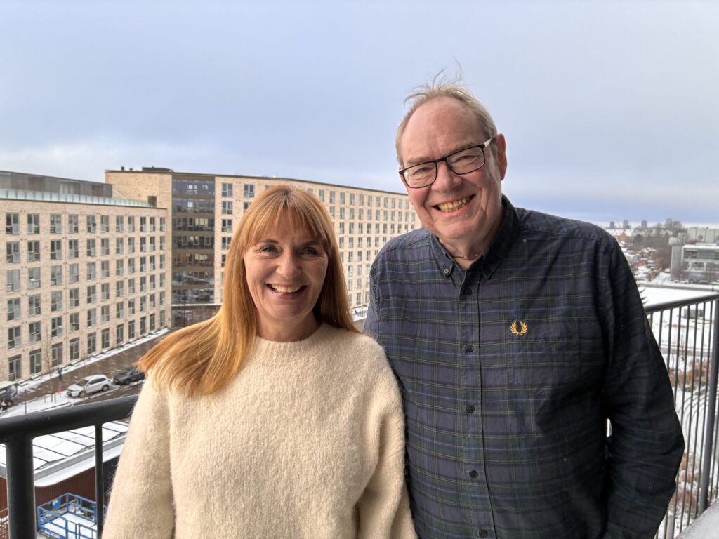 Brian og Susanne smiler, mens de står side om side på en altan med moderne boligblokke og et snedækket bybillede i baggrunden