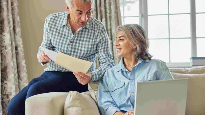 En ældre investor med papirer i hånden taler med en smilende kvinde med en bærbar computer i en sofa i en lys stue med et stort vindue, og begge arbejder på at bevare roen og undgå paniksalg.
