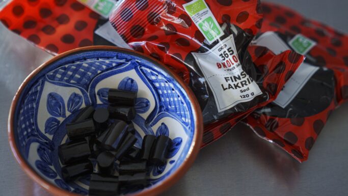 En blå og hvid mønstret skål fyldt med sorte lakridsstykker står foran flere røde og sorte polkaprikkede lakridspakker med påskriften "FIN SALT LAKRIS", en yndlingsgodbid, som mange danskere spiser på trods af den unikke smag.