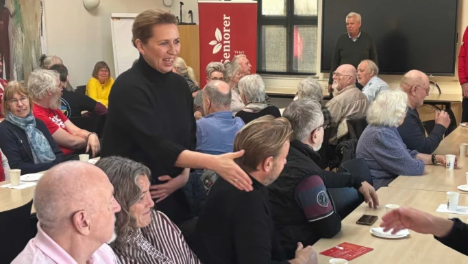 statsminister Mette Frederiksen sort sweater hilser på en siddende mand i et overfyldt lokale med ældre mennesker. Folk taler og smiler og diskuterer pensionsemner over en kop kaffe. I baggrunden ses et rødt banner fra Socialdemokratiet og en mand ved en skærm.