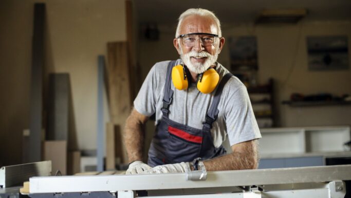 En ældre mand med hvidt hår og skæg smiler, mens han arbejder på et værksted. Han bærer sikkerhedsbriller, gule høreværn om halsen, handsker og overalls. Han er på folkepension og bruger tid på en træbearbejdningsmaskine.