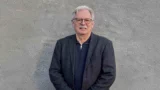 Peter Lautrup står foran en struktureret grå væg iført en mørk blazer over en sort skjorte. Hans neutrale udtryk antyder den ro, der kendetegner en statsminister, der leder en regering.
