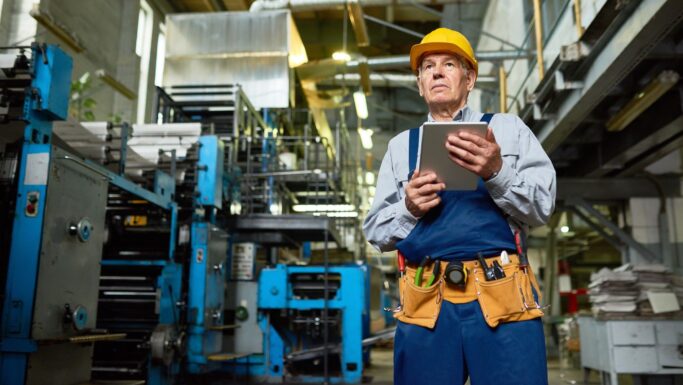 En ældre mand, muligvis en af folkepensionisterne, i gul hjelm og arbejdsdragt står på en fabrik med en tablet i hånden. Han har et værktøjsbælte og er omgivet af store industrielle trykmaskiner.
