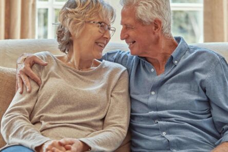 Et ældre par sidder tæt sammen i en sofa og smiler varmt, mens de diskuterer deres pension. Kvinden bærer briller og en beige sweater, mens manden i en blå skjorte ser kærligt på hende. De virker glade og nyder deres pensionisttilværelse i deres kommune.