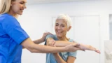 En sundhedsperson hjælper en ældre kvinde med at strække sin arm under en fysioterapisession. Kvinden smiler og ser ud til at have det godt, mens hun arbejder på sine muskler med forsigtig hjælp.