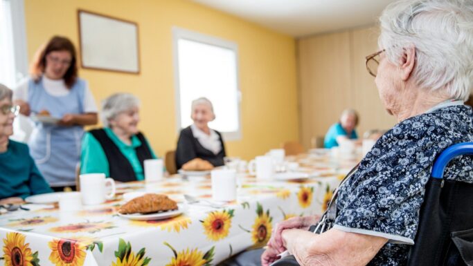 En gruppe ældre kvinder sidder omkring et bord med kaffekopper og kager i et lyst plejehjem og sludrer i et rum med en solsikkedug. En plejer står i nærheden, smilende og med en notesblok i hånden, og sørger for, at de nationale retningslinjer bliver fulgt.