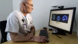 En ældre mandlig læge i hvid kittel undersøger farverige hjernescanninger på en computerskærm ved sit skrivebord ved hjælp af et tastatur og en mus i et veloplyst kontor, hvor han undersøger myter og tidlige tegn på demens.