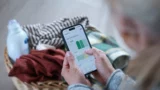 En person holder en smartphone, der viser en graf om gratis strøm, med en vasketøjskurv med tøj og en flaske vaskemiddel i baggrunden.