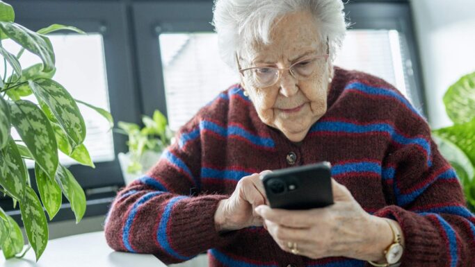 En ældre kvinde med hvidt hår og briller, iført en stribet rødbrun og blå sweater, sidder indendørs og bruger sin smartphone. Hun ser fokuseret ud, måske læser hun om millionbedrageri eller hvordan svindel kan føre til fængsel, omgivet af stueplanter og sollys.