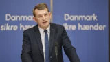 En mand i jakkesæt og briller taler på et podium med en mikrofon. Bag ham er der et blåt bagtæppe med hvid dansk tekst, hvor der står "Danmark sikre hænder." Arrangementet diskuterer emner som folkepension under ledelse af Troels Lund Poulsen.