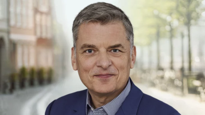 En midaldrende mand med kort, gråt hår, iført blå blazer og ternet skjorte, smiler til kameraet. Det udendørs bymiljø afspejler en følelse af tryghed og frihed med slørede bygninger og træer i baggrunden.