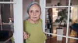 En ældre kvinde med langt gråt hår, iført grøn skjorte og diskrete smykker, står i en døråbning og ser direkte ind i kameraet med et alvorligt udtryk. Baggrunden viser et blødt oplyst rum med en plante.