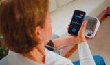 En person bruger en digital blodtryksmåler på armen, mens han holder en smartphone, der viser en AI-svar chatbot-grænseflade, siddende i en sofa i et hjem.