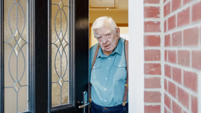 En ældre mand med hvidt hår, iført blågrøn skjorte og seler, står i døråbningen til et hus og kigger ud med et alvorligt udtryk - som en svindler, der er bange for at blive fanget, og hans fortid kaster skygger over murstensvæggen til højre for ham.