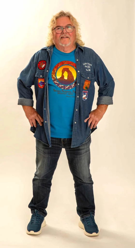 En smilende singler med langt, krøllet blondt hår og briller står med hænderne på hofterne iført jeans, blå sneakers, en blå grafisk T-shirt og en åben denimskjorte prydet med farverige lapper - en ægte Hotel Romantik-stemning.